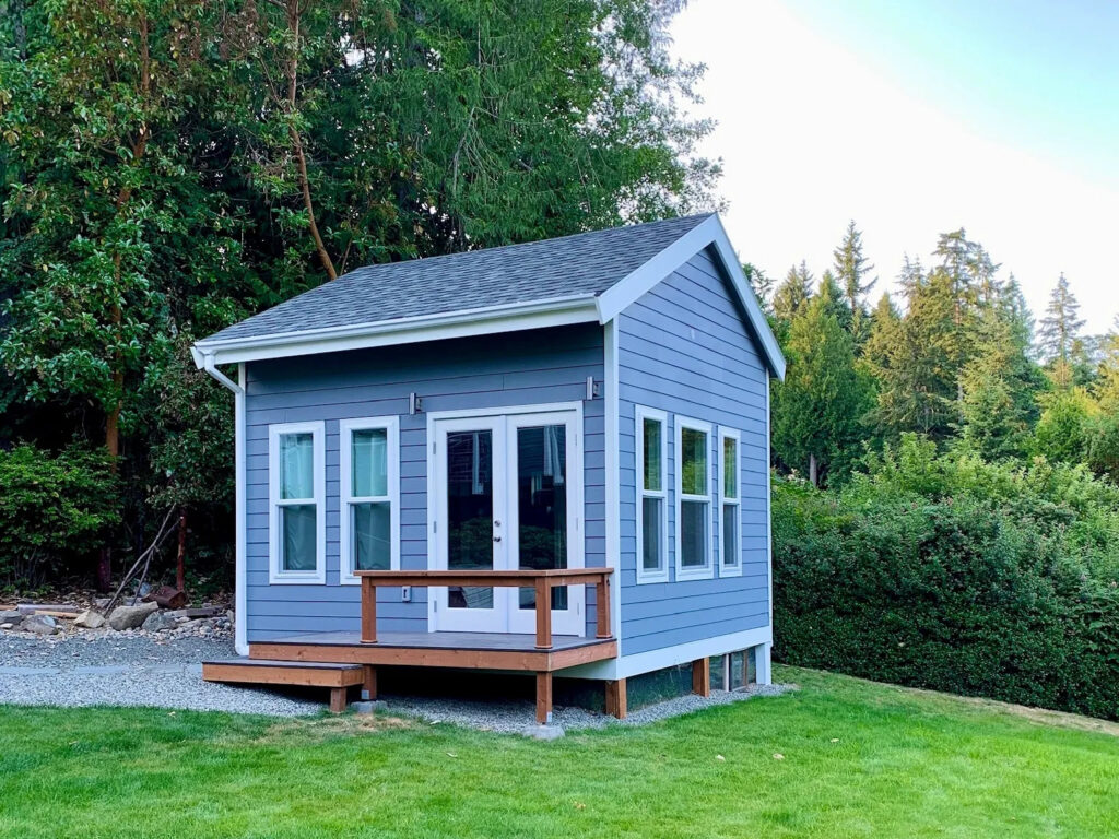 image of ADU with gray siding, white trim, glass french doors, wooden decking, and evergreen trees and shrubs in the background.