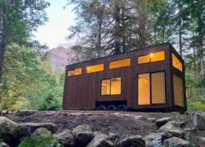tiny house on wheels in forest setting. exterior is brown Shou Sugi Ban siding. interior lights give a golden glow.