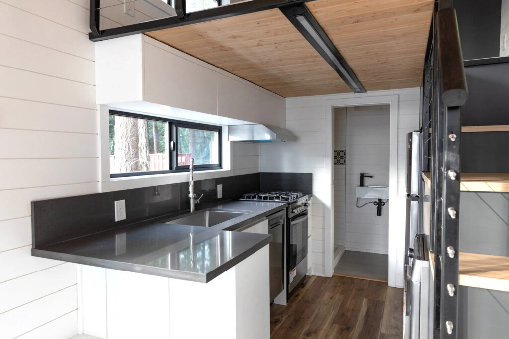 interior image of tiny house kitchen and stairs. lap siding, lvp flooring, quartz counters, and entrace to bathroom.
