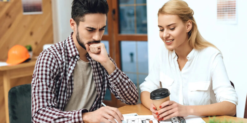 two people sitting at table looking at building plans. construction hat in background.